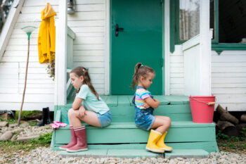 Siblings sitting  back to back avoid talking . Kids  quarreling and sulking  each other on porch at backyard against house. Life in the suburbs.